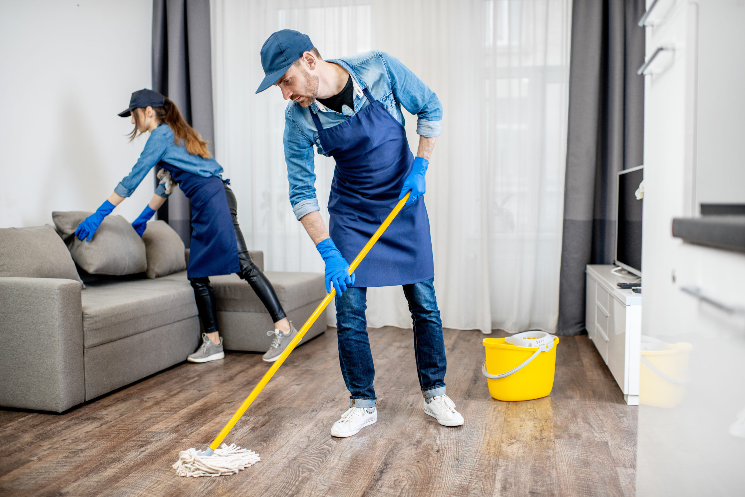 Professional cleaners in the apaartment Professional cleaners in blue uniform washing floor and wiping dust from the furniture in the living room of the apartment. Cleaning service concept